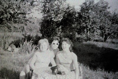 Two women seated on a small hill with tall grass and dense foliage in the background. Both appear to be wearing swimwear. App...