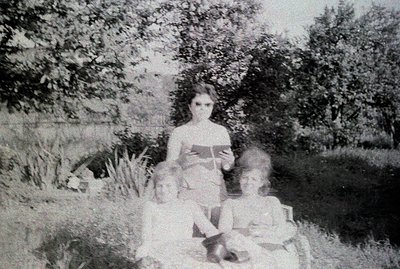 Black and white photograph showing a woman seated with two young children. The woman holds a book, positioned outdoors in a g...