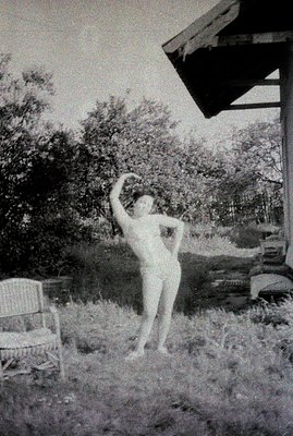 A woman stands outdoors, partially sheltered by a cantilevered roof, in a pose suggestive of ballet or dance. Lush greenery f...