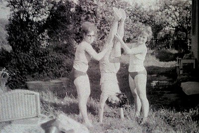 Three young women playfully balance two others upside down, suspended by their ankles. The outdoor scene features a grassy la...