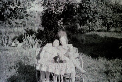 Three young girls in dresses gather near a small, wrought-iron table and chairs within a grassy, wooded area. The image has v...