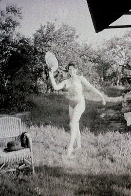 A woman in swimwear playfully tosses a frisbee in a grassy yard, likely a backyard. A wicker chair with a blanket rests nearb...