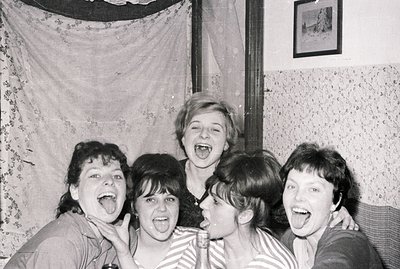 Group portrait of five young women, displaying playful expressions, likely friends, indoors. Wallpaper, floral curtains, and ...
