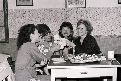 Four women toasting with mugs in a vintage cafe setting, likely 1960s-70s. Decorative wallpaper, a small table with pastries,...