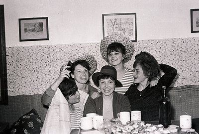 Five young women playfully pose, adorned with various straw hats. A table laden with cups, a wine bottle, and a plate of mars...