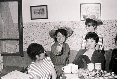 Four women socialize in a richly patterned interior; two wear striped tops & straw hats. A table with teacups & pastries sits...