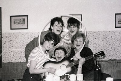 A group of five young adults playfully pose for a casual portrait. Seated amongst teacups, one plays a guitar, another dons a...