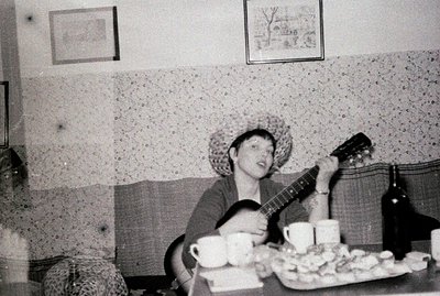 A young person seated at a table, playing an acoustic guitar. Visible are several cups and a platter of what appears to be fr...