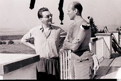 Two men converse on a balcony overlooking a forested landscape. One smokes, both wear glasses. Casual summer attire suggests ...