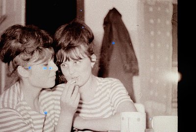 Two young women, striped tops, close proximity. One gently touches the other's face. Interior scene, likely a home, with a da...