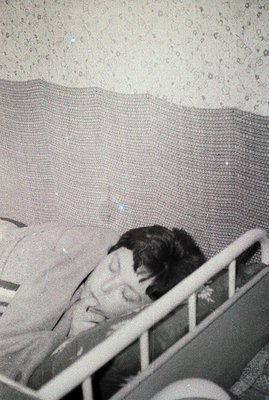 A young child sleeps in an iron crib with a textured, woven blanket. The grainy image suggests a vintage photograph, potentia...