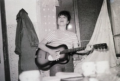 A young woman stands playing an acoustic guitar in what appears to be a sparsely furnished room. She wears a striped shirt an...