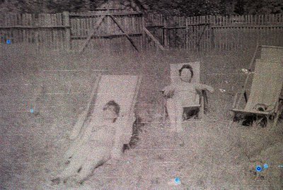 An outdoor scene depicts three figures, likely children, standing in what appears to be a garden or yard. The image quality i...