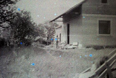 A grainy, black-and-white photograph captures a simple, single-story building with a low-pitched roof and a visible window. T...