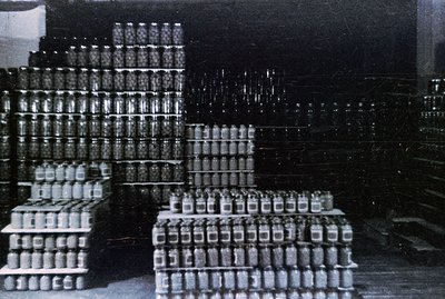 Stacked crates overflow with glass jars, likely canned goods, within a dark warehouse or factory setting. The monochrome imag...