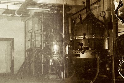 Industrial interior featuring large, complex machinery—likely processing or refining equipment. Visible piping, gauges, and m...
