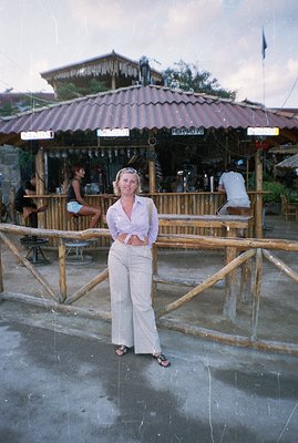 A woman stands near a beachside bar, posed for a portrait. She wears a cropped floral shirt and wide-leg trousers; sandals su...
