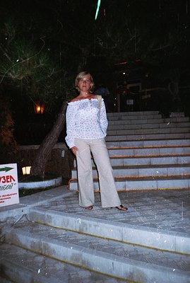 A woman stands on stone steps at night, wearing a patterned off-the-shoulder blouse and flowing wide-leg trousers. Sign visib...
