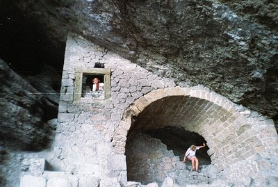 Stone archway & window integrated into a rugged, dark rock face. A figure sits within the arch, another observes from above. ...