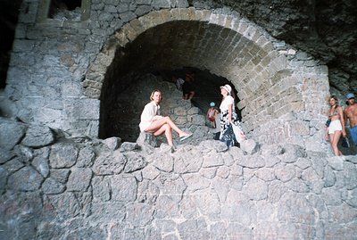 Three young women are seen within a stone archway, seemingly part of a coastal structure. One woman sits, while the others st...