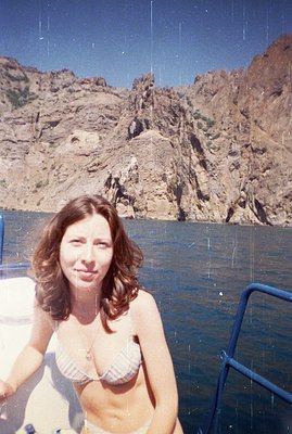 A woman with shoulder-length dark hair sits on a boat, wearing a patterned bikini and a delicate necklace. Dramatic, rugged c...