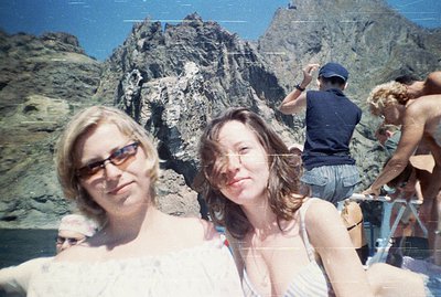 Two young women, one wearing sunglasses, pose outdoors with a rugged, rocky mountain backdrop. Several other figures are part...