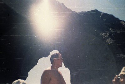 A man in a short-sleeved shirt stands in profile, backlit by bright sunlight over a mountainous, rocky landscape. Likely a to...