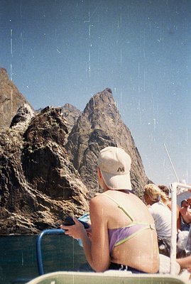 A person in a purple bikini and cap is looking at a phone on a boat, with a dramatic, rocky mountain range visible beyond. Th...