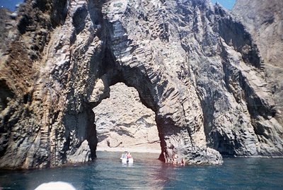 Dramatic coastal rock formation with a natural archway. Boats and figures visible near the shore within the arch. Heavily tex...