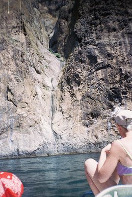 A person in swimwear sits on an inflatable raft on a turquoise river, facing a sheer, rocky cliff face with sparse vegetation...