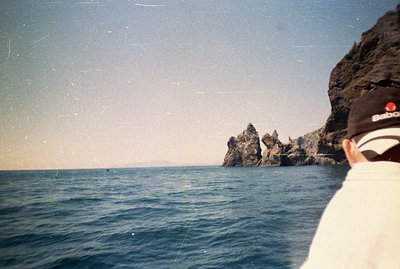 View from the sea towards rocky cliffs & a distant shoreline. A person wearing a Babolat visor and dark clothing occupies the...