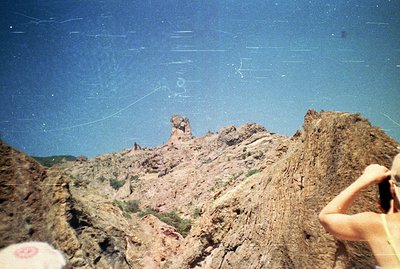 Striking, eroded sandstone rock formations dominate the scene against a bright blue sky. A person’s arm and hand, holding sun...