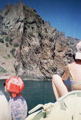 A view of layered rock formations reflected in still, turquoise water. Two figures are partially visible, wearing a red banda...