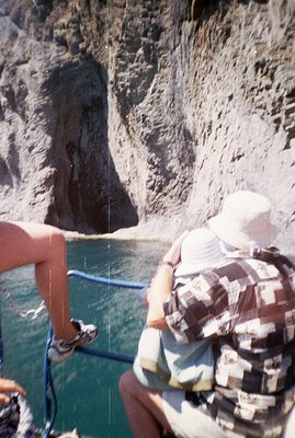 A person wearing a white bucket hat and plaid shirt poses on a boat, overlooking turquoise water and a steep, rocky cliff fac...