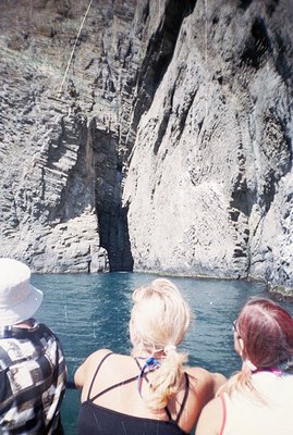 A view from a boat, observing towering coastal cliffs with layered rock formations. Two women with blonde and auburn hair are...