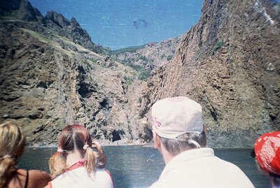 The photograph depicts a family viewing towering, rocky cliffs rising from a body of water, likely a river or lake. Three ind...