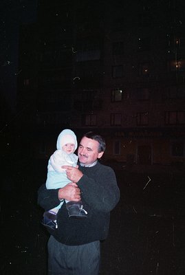 A man carries a baby bundled in a white snowsuit in front of a building with illuminated windows. The building features archi...
