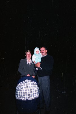A family portrait: a smiling baby in a white cap is held by a man in a dark sweater, standing next to a woman in a gray plaid...