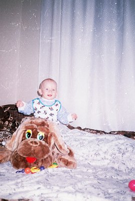 A baby, dressed in a patterned onesie, sits atop a large plush dog toy surrounded by colorful balls. The image appears to be ...