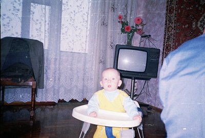 Infant sits in a plastic highchair, gazing directly at the camera. Room features floral curtains, rug, & a vintage television...