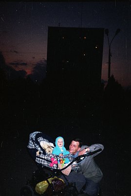 Man pushing stroller with baby in a blue onesie. Stark, Brutalist apartment block dominates the background with lit windows. ...