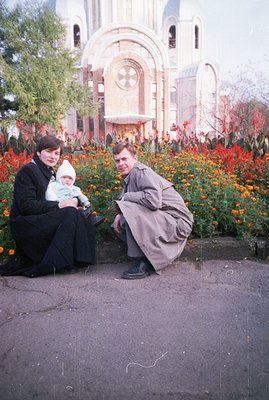 A family of three poses before a grand, ornate Orthodox church building. The mother, in a dark dress, holds a baby wearing a ...