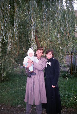 A man in a trench coat holds a baby bundled in white. A woman in a long black coat stands beside them, both facing the camera...