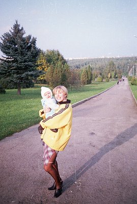 A woman in a bright yellow jacket carries a baby in a sling along a paved path. Lush greenery & trees frame the scene. Appear...