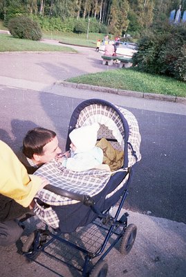 A man in a yellow shirt leans over a baby in a vintage pram on a paved pathway. Trees and a grassy bank form the backdrop. Th...