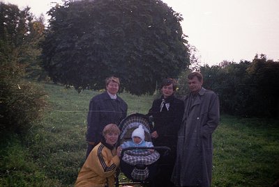 A family portrait, likely taken in the 1970s. Four people pose outdoors near a large evergreen shrub. A baby is in a stroller...