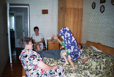 A family scene: A young boy playfully jumps onto a man reclining on a patterned bedspread, while a woman watches with amuseme...