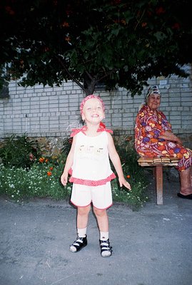 A young child in a white and red patterned dress stands outdoors, facing the camera with a wide grin. An elderly woman sits o...