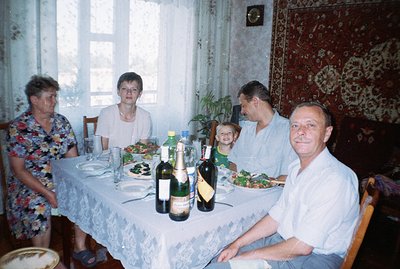 A family gathers around a table laden with food and drinks. Visible are several bottles of wine, salads, and bread. The room ...