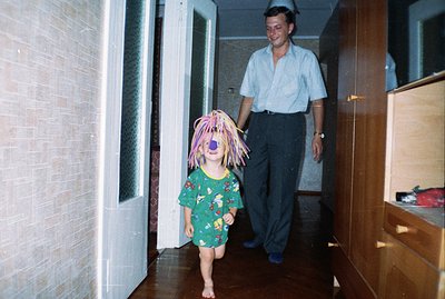 A barefoot child in patterned pajamas and a bright orange wig playfully runs toward the camera in a hallway. An adult man, we...